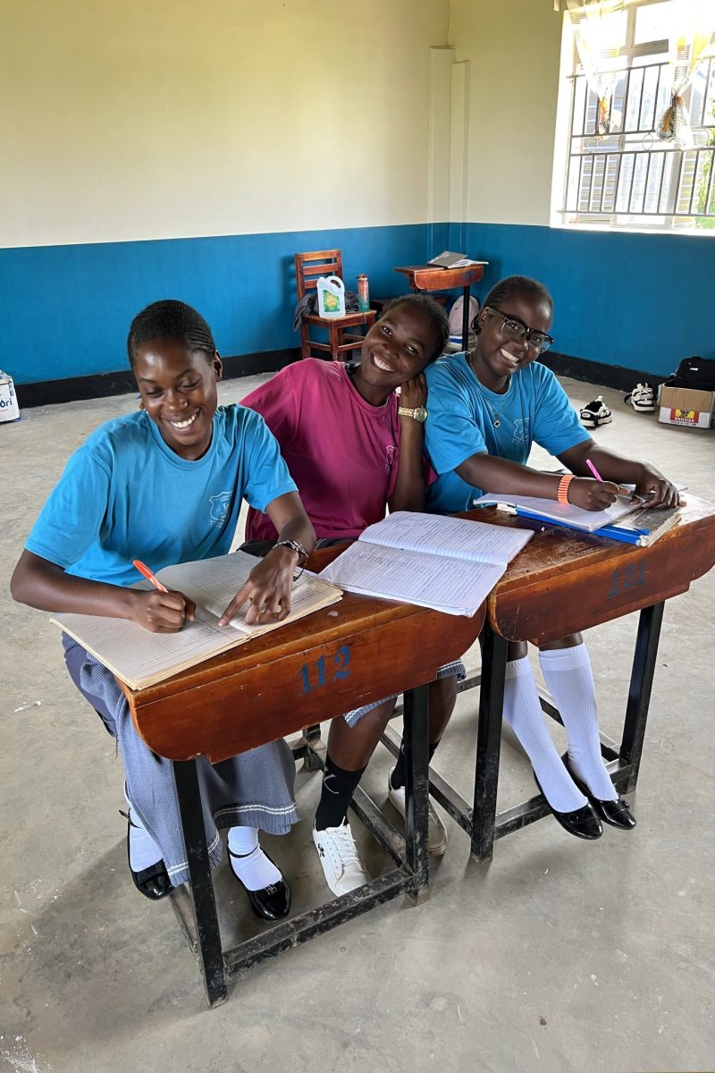 Three senior high girls, seated together at desks, look up from their school work to smile fro the camera.