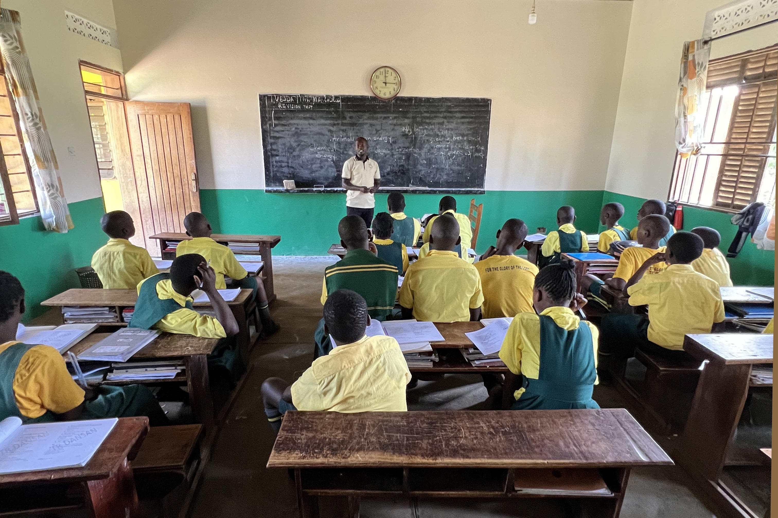 Primary 7 students are sitting in their desks, looking at their teacher and listening attentively to him.a